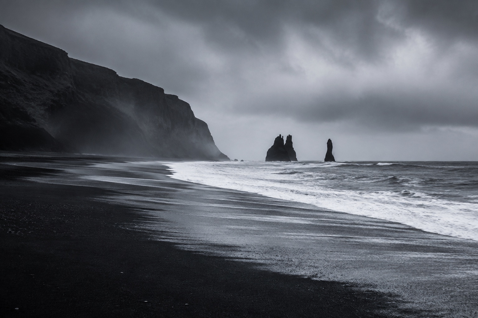 Waves crossing a black sand beach beneath cliffs and a low grey sky