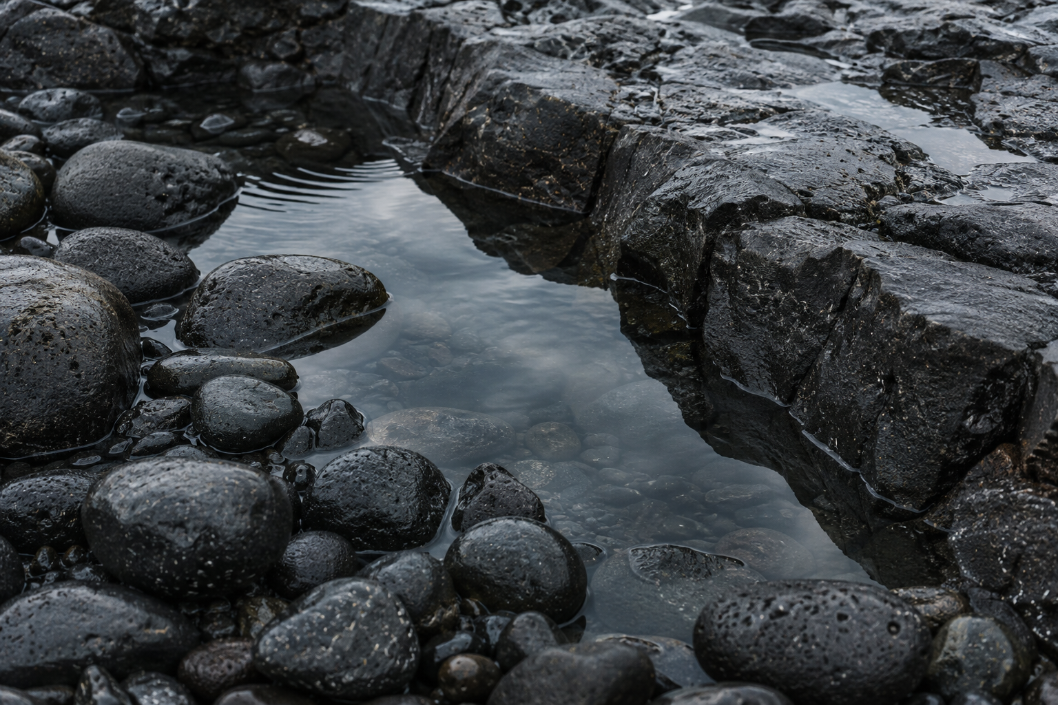 Rainwater reflecting a grey sky between rounded volcanic pebbles and wet basalt